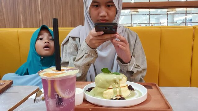 Samarinda, Indonesia. January 23th 2023. Happy Asian muslim teenage girl sitting in cafe and taking picture of her food with smart phone before eat