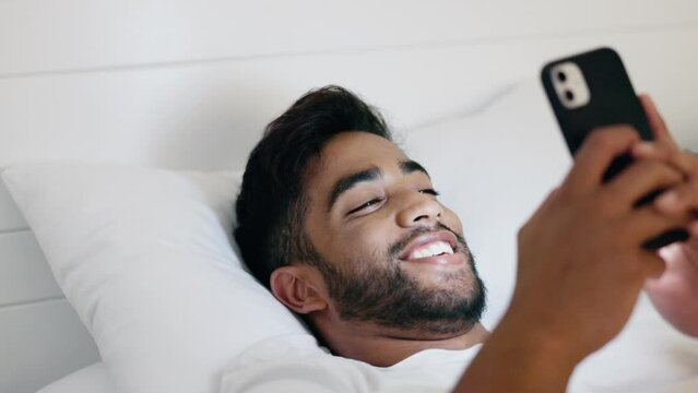 Man, phone and smile lying in bed for social media, chatting or texting while relaxing at home. Happy relaxed male smiling for morning entertainment on smartphone in bedroom browsing app or streaming