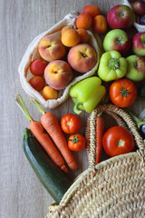 Straw bag and reusable fabric bags filled with various healthy fruit and vegetables. Wooden background, top view.