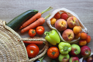 Straw bag and reusable fabric bags filled with various healthy fruit and vegetables. Wooden background, top view.