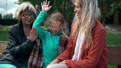 Smiling happy senior and adult women and girl stacking hands sitting on bench in spring summer park. Positive Caucasian grandmother mother and daughter enjoying leisure on windy day
