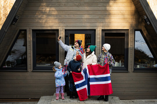 Portrait Of Family With Kids Outside Cabin House Holding Norway Flags And Making Selfie On Phone. Scandinavian Culture, Norwegian People.