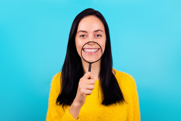 Photo of nice gorgeous adorable girl wear yellow sweater hold magnifying glass on teeth toothy beaming isolated on blue color background
