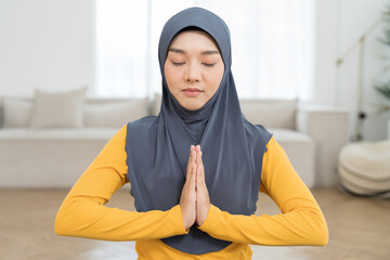 Asian Young Muslim woman doing meditate yoga asana on roll mat with eyes closed in home. Girl practicing doing yoga indoor