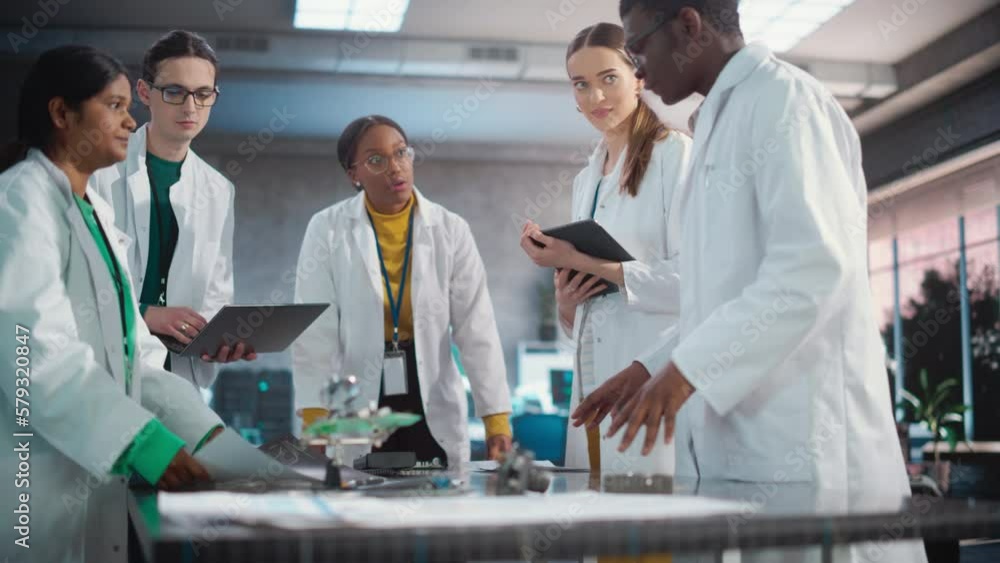 Poster Engineers In Lab Coats Using Laptop Computer and Tablet, Brainstorming Ideas For Electronic Components. Diverse Team of Industrial Robotics Specialists Gathered Around a Table to Discuss a Blueprint