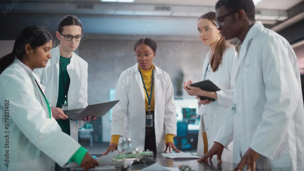 Poster Engineers In Lab Coats Using Laptop Computer and Tablet, Brainstorming Ideas For Electronic Components. Diverse Team of Industrial Robotics Specialists Gathered Around a Table to Discuss a Blueprint