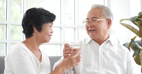 Fototapeta premium Asian elderly couple couple sitting on sofa at home drink water for health smiling healthy senior retired grandparents husband and wife