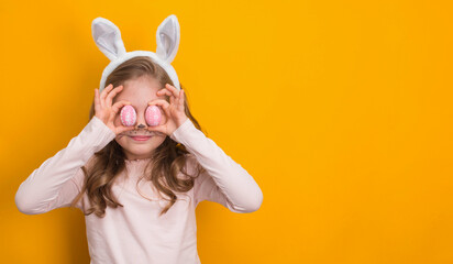 Funny girl with Easter bunny ears holding decorated easter eggs on yellow background with copy-space.