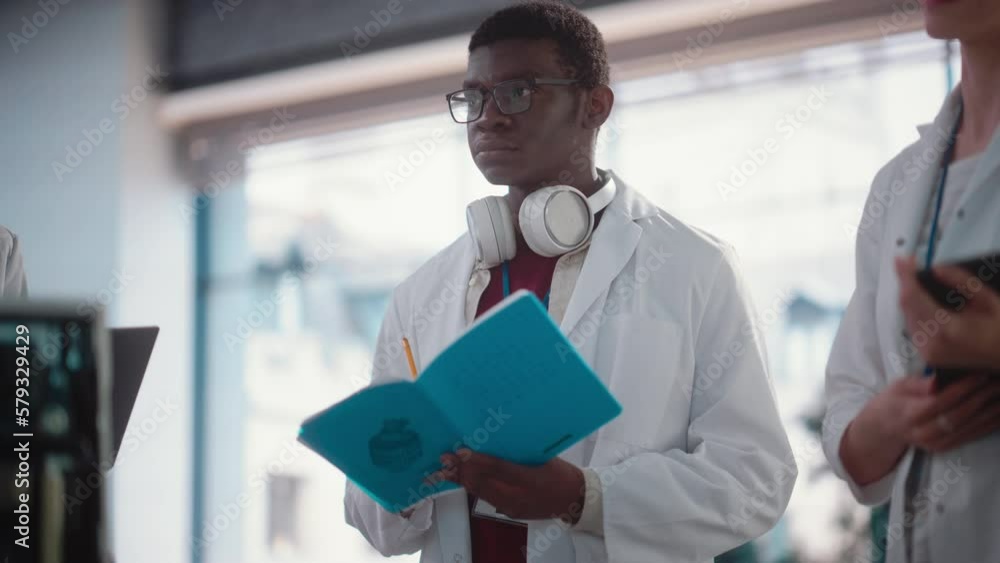 Poster Slow Motion Portrait of Young Black Male Engineering Student Asking Questions and Taking Notes in a Laboratory. Lab Assistant in Meeting Showing Initiative and Giving Ideas, Brainstorming with Team