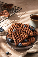 Heart form chocolate waffle cookies in brown ceramic plate decorated with berries with tea cup on beige background. Sunny morning breakfast with hursh shadows