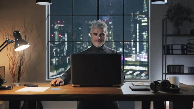 Mature male at the desk using computer at night stop working closes laptop and stretching,relaxing,modern home office with window city view in the background
