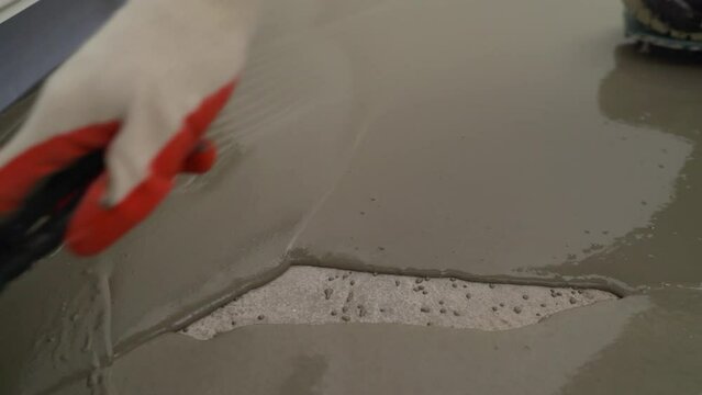 Repairman in protective gloves and uniform using a metal spatula to level a liquid self-levelling floor screed. A worker pours liquid cement mortar at a construction site. The master using a toothed t