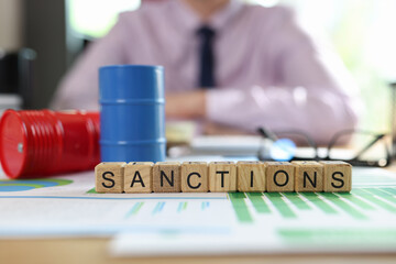 Wooden cubes with word sanction, oil drums and documents on table and msnager in background.