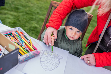 little boy looking serious drawing an Easter egg at an Easter fair, a grandmother helps him hold a...