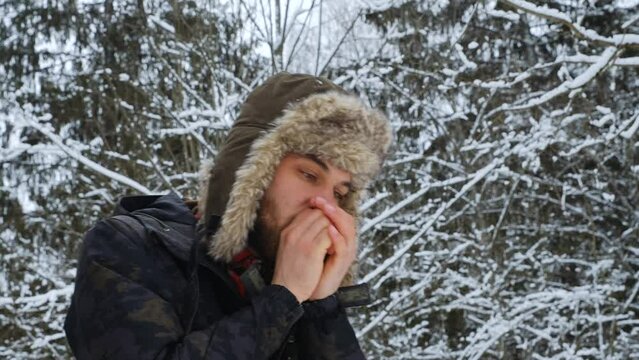 Man froze on walk outside in winter and trying to warm up, warming hands with breath. Concept of problem and peculiarities nature of North. Guy in hat with earflaps in snow park.