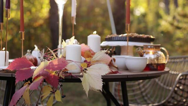 Cozy autumn picnic in the park. Close up of table setting with white plates, cutlery, glass vase with colourful wildflowers, homemade apple pie, maple leaves, burning candles on wooden table outdoors