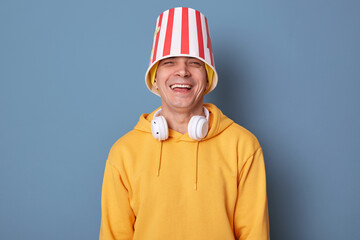 Portrait of happy overjoyed man wearing beanie hat and casual yellow hoodie posing against blue wall, standing with popcorn bucket on head, laughing out loud, having fun in cinema