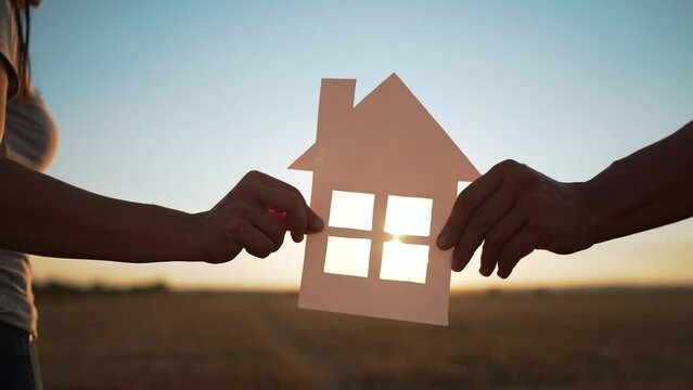 Happy family. People hands hold a paper house together. House made of paper is symbol of family happiness, mortgage protection, ecology. Sun shines through the window of house in nature at sunset