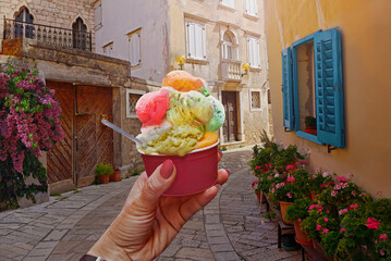 Woman hand hold  beautiful bright sweet ice - cream cone with different flavors  held in hand on the background of old street  in  Porec,Croatia. Traveling concept background - 579355014