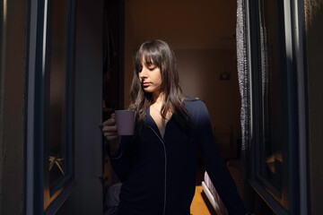 young woman drinking coffee or tea in pajamas at the window at home