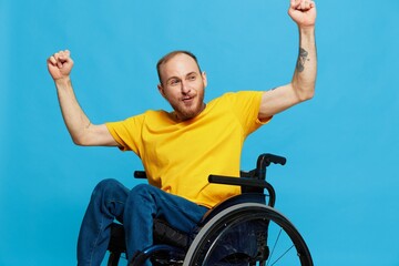 A man in a wheelchair happiness raised his hands up in a t-shirt with tattoos on his hands sits on a blue studio background, a full life, a real person