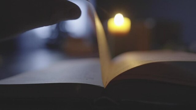 A man's hand turns the page and smooths them in the foreground. A defocused candle stands in the foreground. High quality 4k footage
