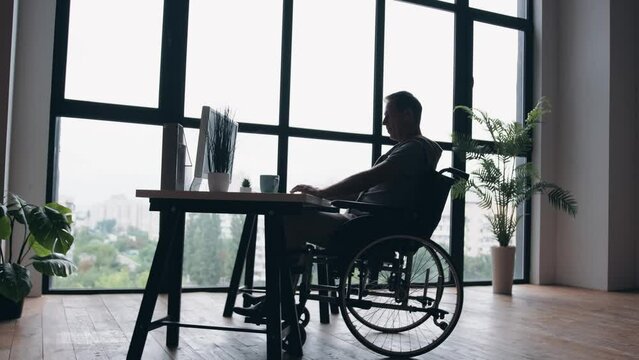 An elderly man in a wheelchair works in the office at the computer. Silhouette of a disabled man