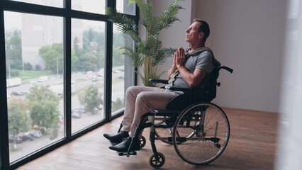 An elderly man in a wheelchair prays near the window. Disabled man praying to god