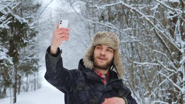 Young Caucasian male blogger in fluffy hat with earflaps stands in middle of winter forest and takes selfie video on phone. Friendly guy talking on video link outside and smiling. Slow motion footage.