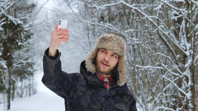 Young Caucasian male blogger in fluffy hat with earflaps stands in middle of winter forest and takes selfie video on phone. Friendly guy talking on video link outside and smiling. Slow motion footage.