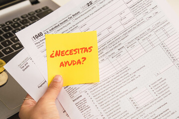 Hands holding a help text in spanish on sticky notes with 1040 tax forms , United States tax form, a laptop on a desk near some bitcoins. US individual income tax return. 