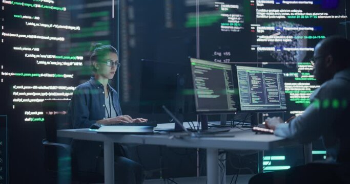 Young Man and Woman Working on Desktop Computers, Surrounded by Big Screens Displaying Code Lines. Professional Computer Engineers Developping System Control and Monitoring Telecommunications