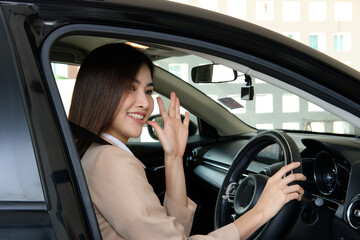 Fototapeta na wymiar Young businesswoman behind the driver's seat talking on the phone while stopping and waving hello through the window of her car