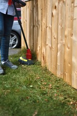 Female cutting mowing and edging grass lawn beside a wooden fence with an electric trimmer