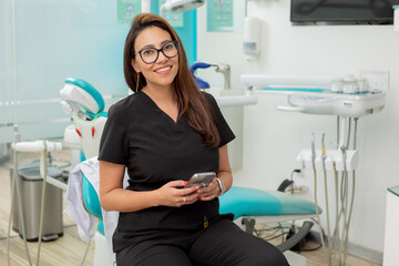 Young female dentist looking at camera smiling, sitting in her office with a cell phone in her hand
