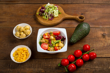 Vegetable salad with champignon corn and cherry tomato with avocado in a white plate on a wooden table next to the ingredients.