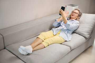 Elderly lady in good mood lies on sofa at home