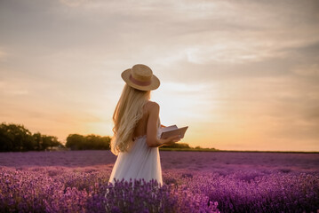 Young beautiful woman in white dress and hat is walking in a lavender field and reading a book.