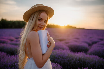 Young beautiful woman in white dress and hat is walking in a lavender field and reading a book.