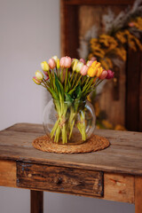 Fresh spring colorful bouquet of tulips in a vase, standing on a wooden table in a warm, cozy living room. Festive flowers for a gift. Mockup for a greeting card.