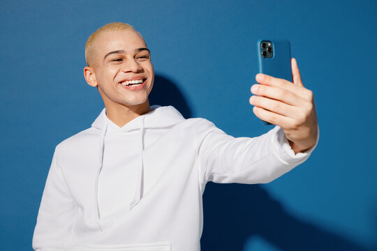 Young dyed blond man of African American ethnicity wear white hoody do selfie shot on mobile cell phone post photo on social network isolated on plain dark royal navy blue background studio portrait.