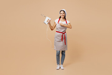 Full body young fun housewife housekeeper chef baker latin woman wear striped apron toque hat hold in hand show point finger on mixer isolated on plain pastel light beige background Cook food concept