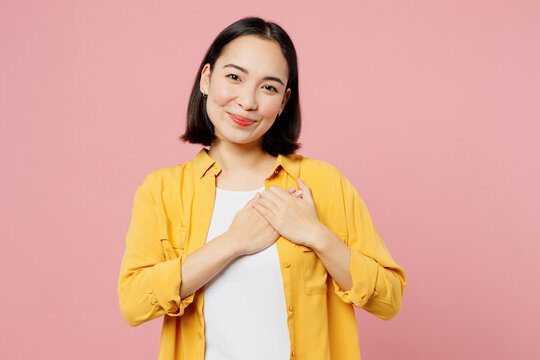 Young grateful smiling thankful woman of Asian ethnicity wears yellow shirt white t-shirt put folded hands on heart isolated on plain pastel light pink background studio portrait. Lifestyle concept.