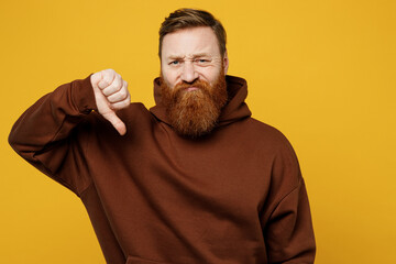 Young dissatisfied displeased sad upset redhead caucasian man wearing brown hoody casual clothes show thumb down dislike gesture isolated on plain yellow background studio portrait. Lifestyle concept.