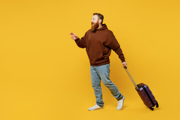 Traveler fun redhead man wear brown hoody casual hold suitcase pointing aside isolated on plain...