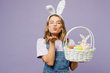 Young cute fun woman wearing casual clothes bunny rabbit ears hold wicker basket colorful eggs blow send air kiss wink blink eye isolated on plain pastel purple background studio Happy Easter concept.