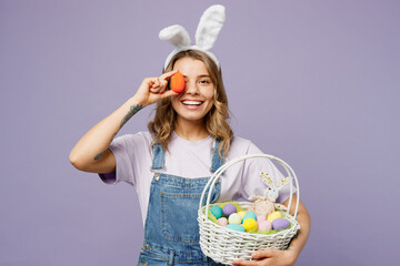 Young smiling fun woman wearing casual clothes bunny rabbit ears hold wicker basket cover eye with colorful violet egg isolated on plain pastel purple background studio portrait. Happy Easter concept.