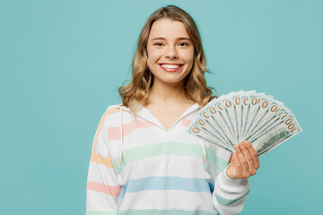 Young smiling fun blonde woman wear striped hoody hold in hand fan of cash money in dollar banknotes look camera isolated on plain pastel light blue cyan background studio portrait. Lifestyle concept.