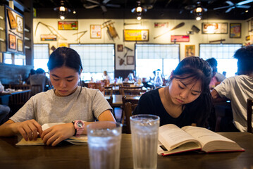 Two young asian girls read books at a restaurant table while their food arrive