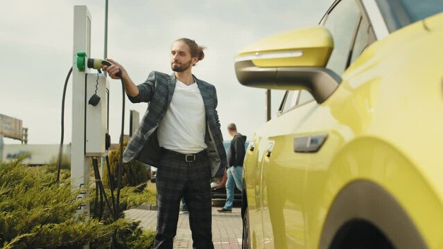 Eco-friendly modern green transport. Hipster fashionable man near electric car. Man charging electric car at a public charging station. Electric vehicle charging port plugging in EV modern car